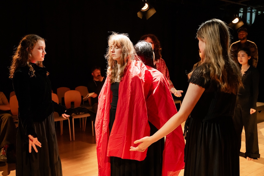 Two actors wearing bright red capes stand with their backs to each other while other actors wearing all black stand in a circle around them.